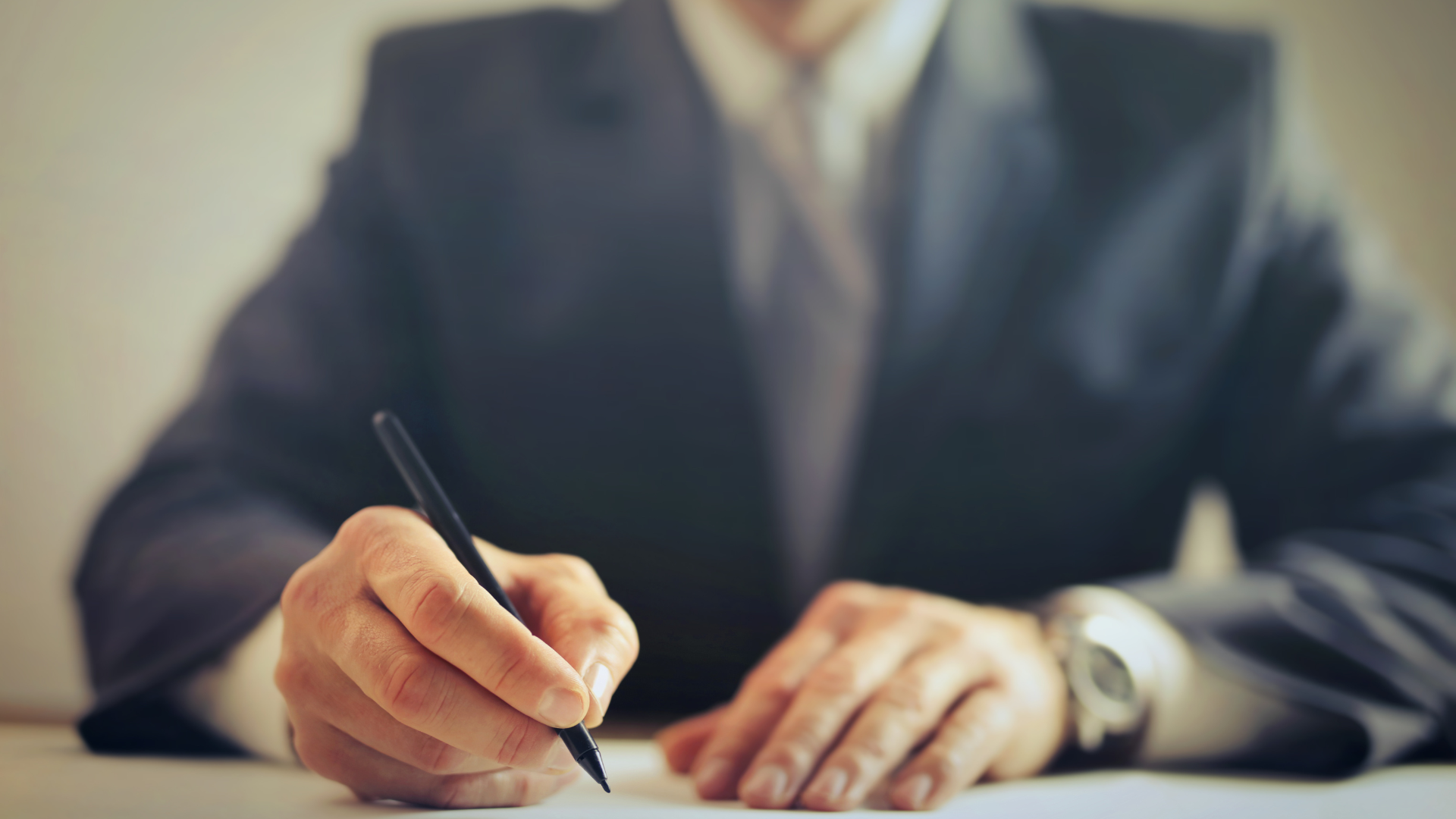 Person in suit writing with a pen on paper, close-up shot of hands and torso.