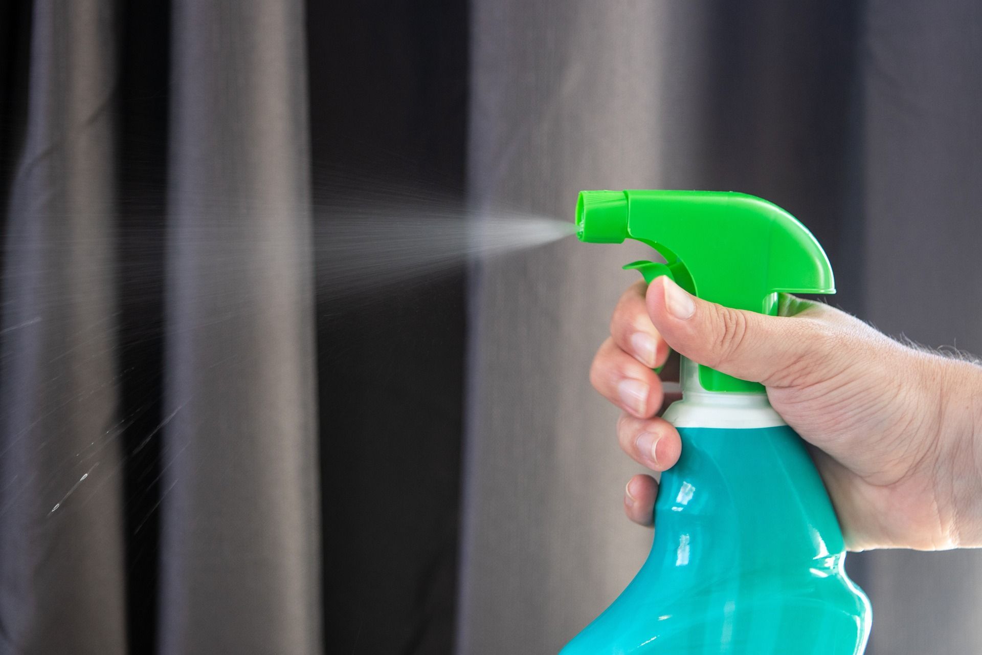 Hand spraying teal spray bottle towards grey curtains.