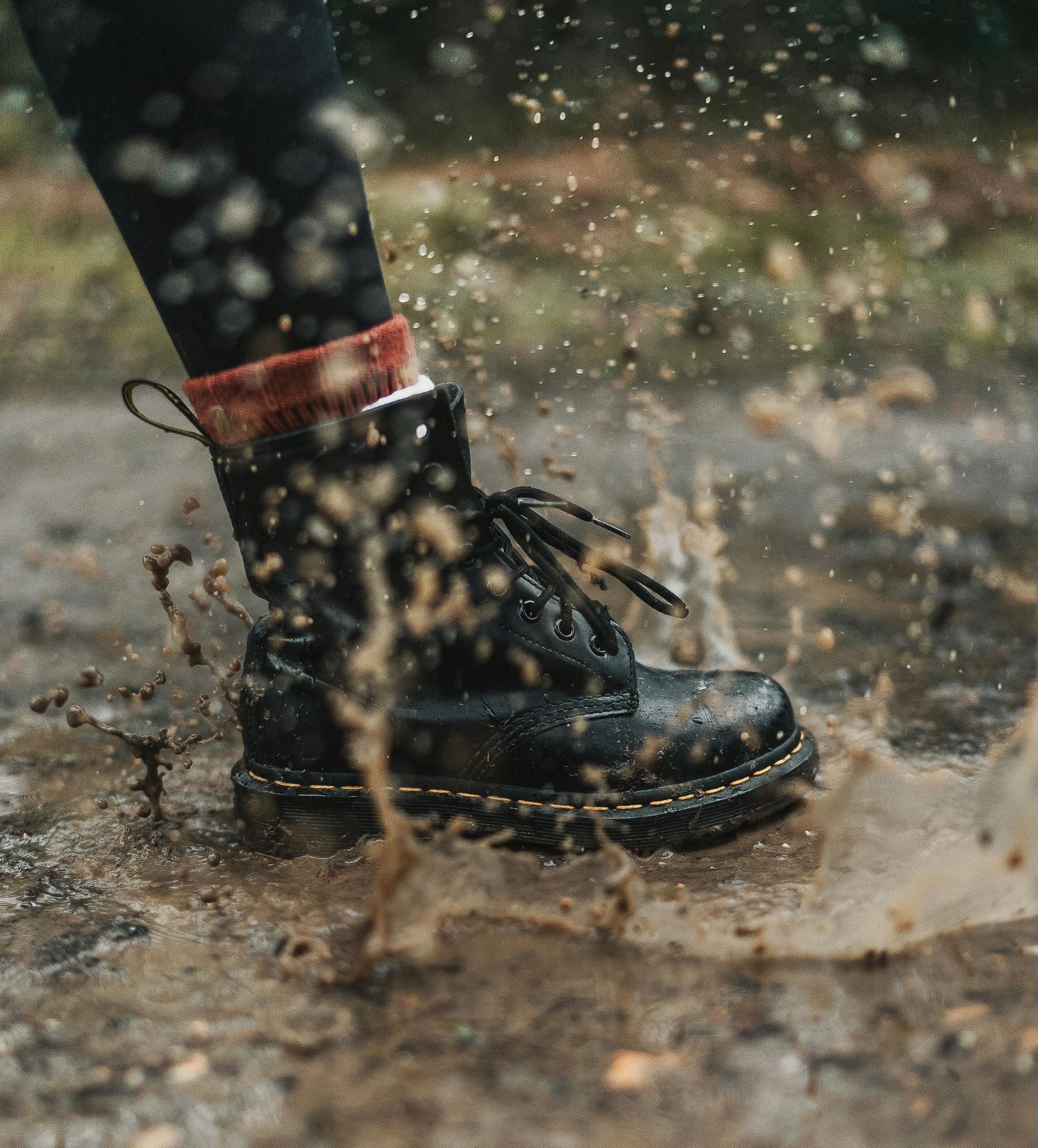 A person wearing black boots is splashing in a muddy puddle.