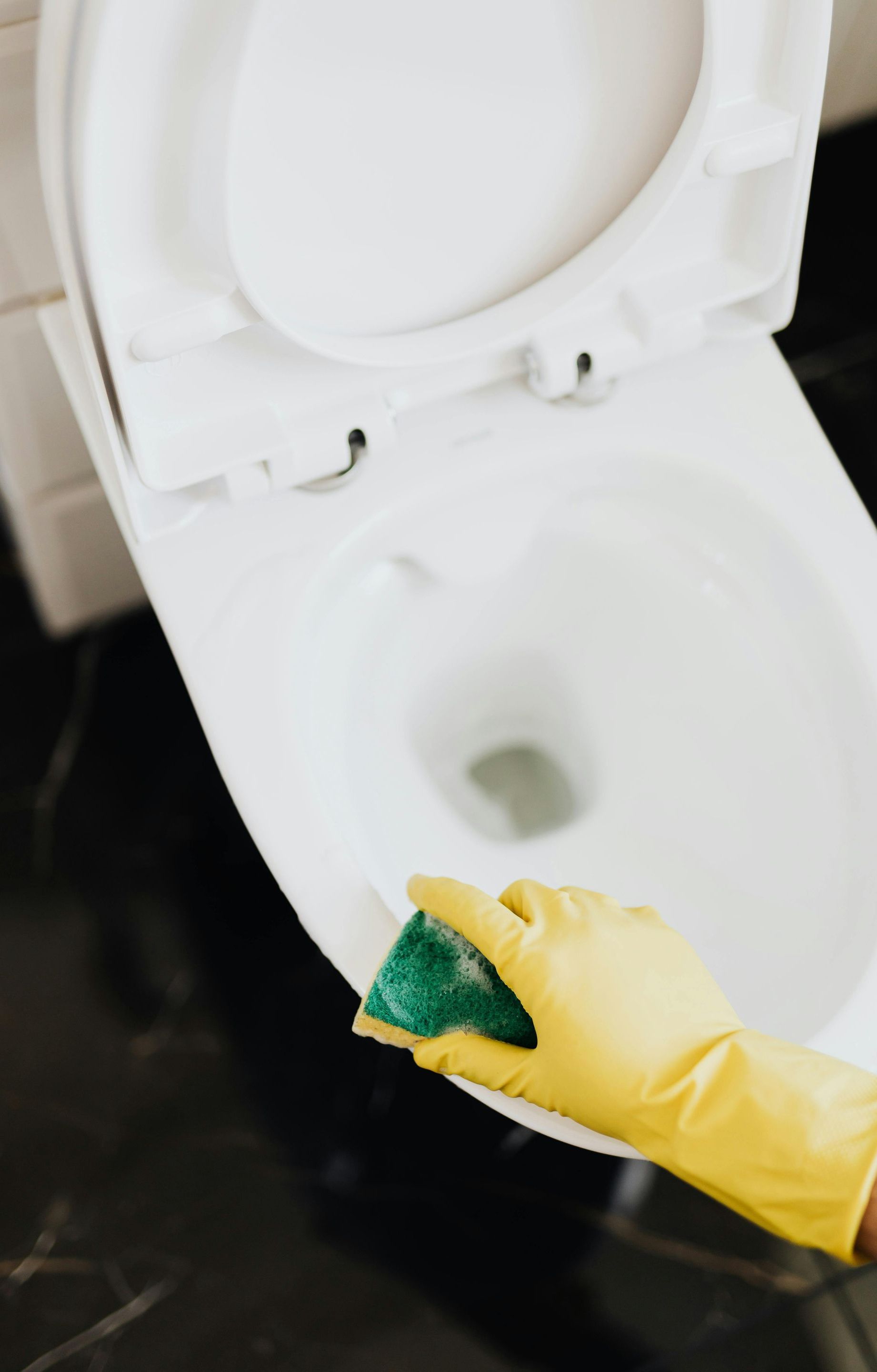 A person wearing yellow gloves is cleaning a toilet with a green sponge.