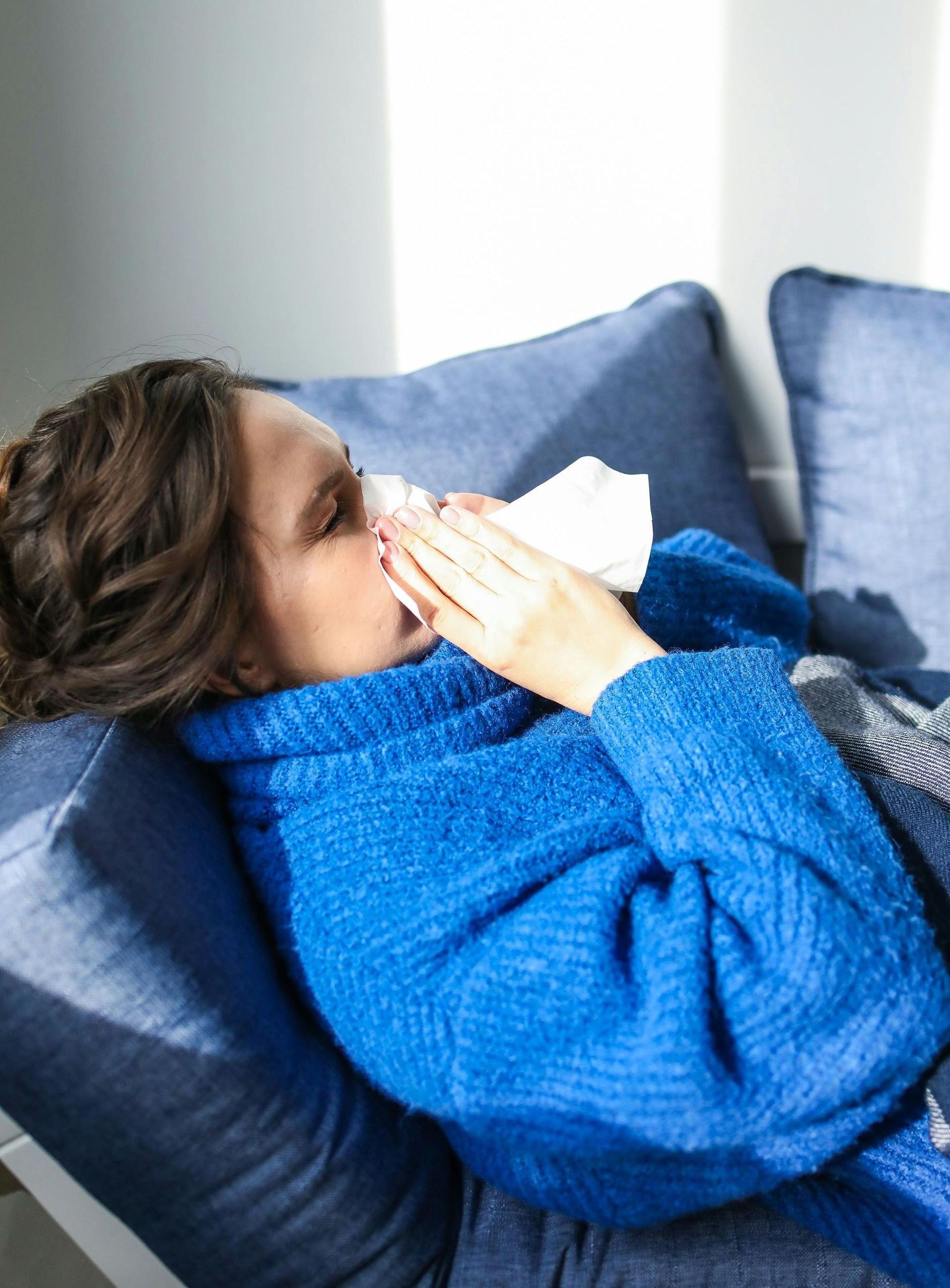 A woman is laying on a couch blowing her nose with a napkin.