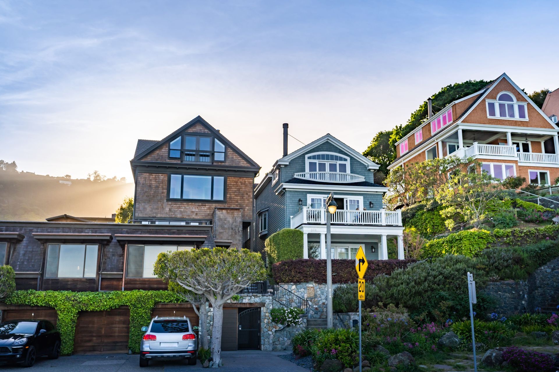 Three multi-story residential houses sit on a hillside with lush greenery, stone walls, and parked cars below.