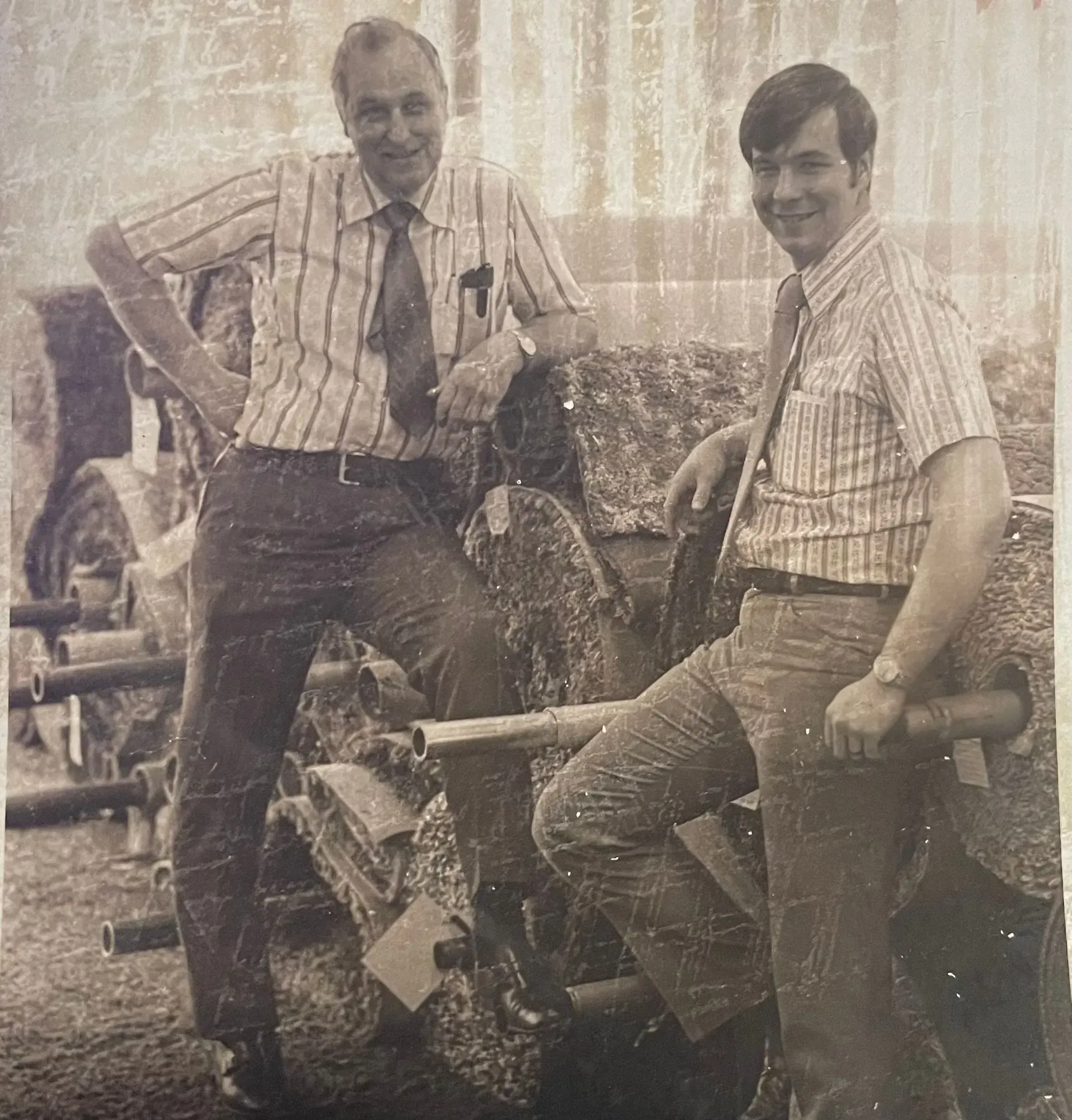 Two men pose beside a muddy tractor, smiling outdoors in a sunlit field.