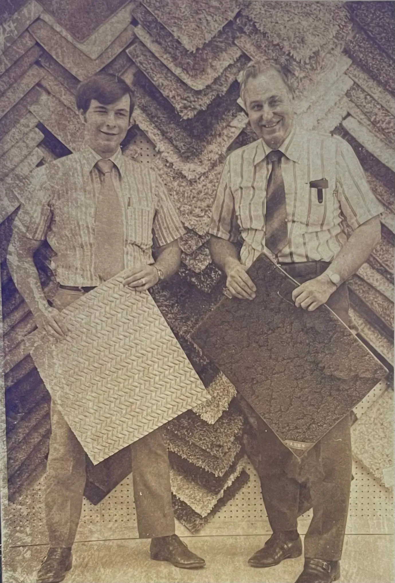 Two men standing outdoors holding large patterned fabric samples against a chevron wall