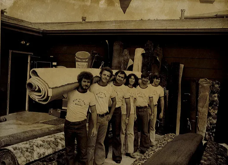 Workers stand in a room holding a large rolled carpet or rug.