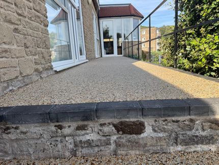 Stone house ramp with gravel surface leading to a glass door and patio, bordered by black railings and greenery