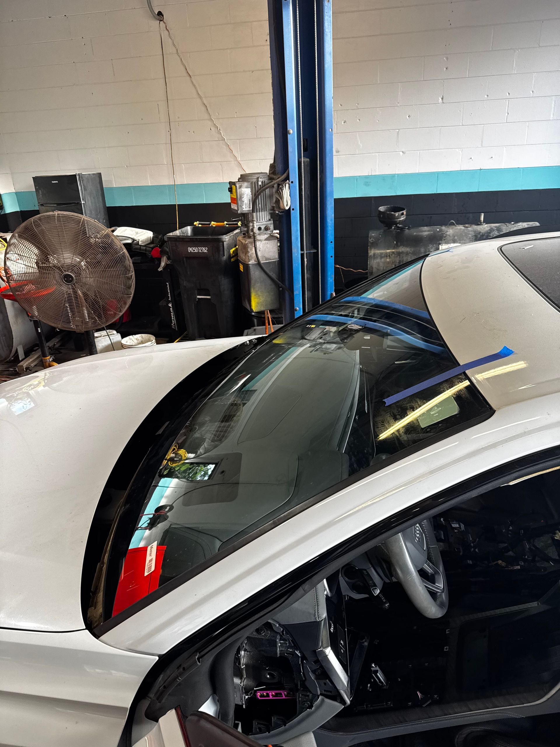 Windshield Replacement: White sports car in an auto shop with the driver door open, next to a large fan and lift posts.