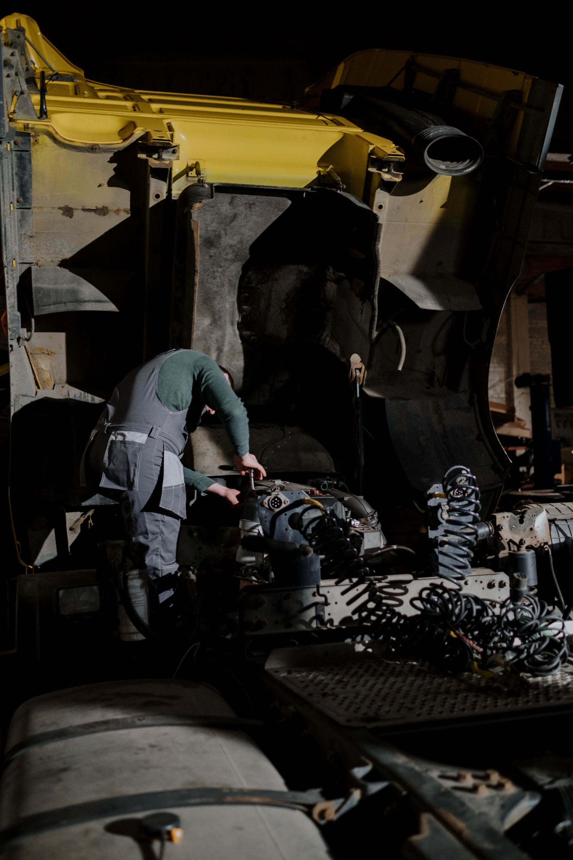 Worker inspecting engine parts in a dim industrial workshop under a yellow overhead machine