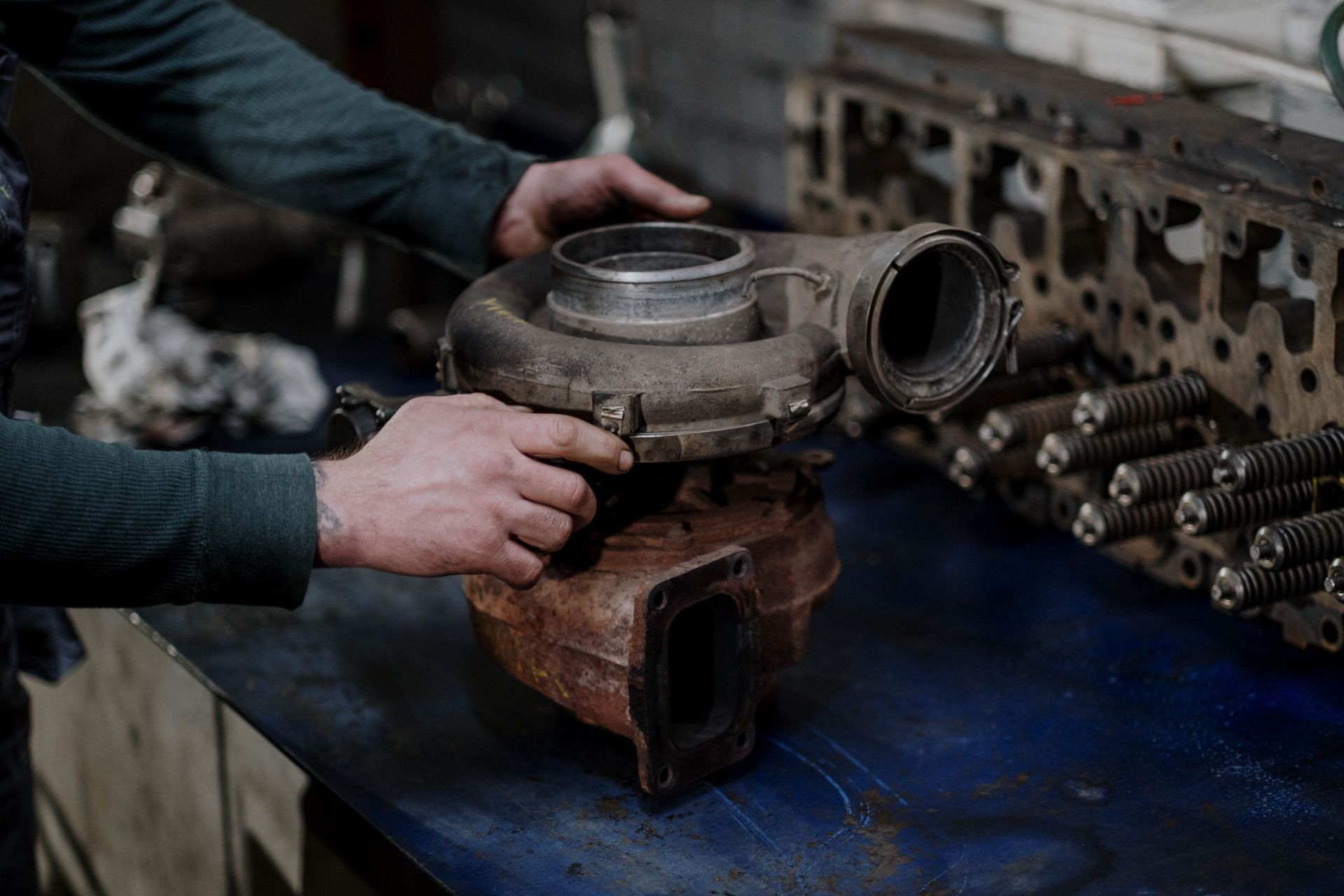 Hands inspecting a rusty turbocharger on a workbench in an auto repair shop