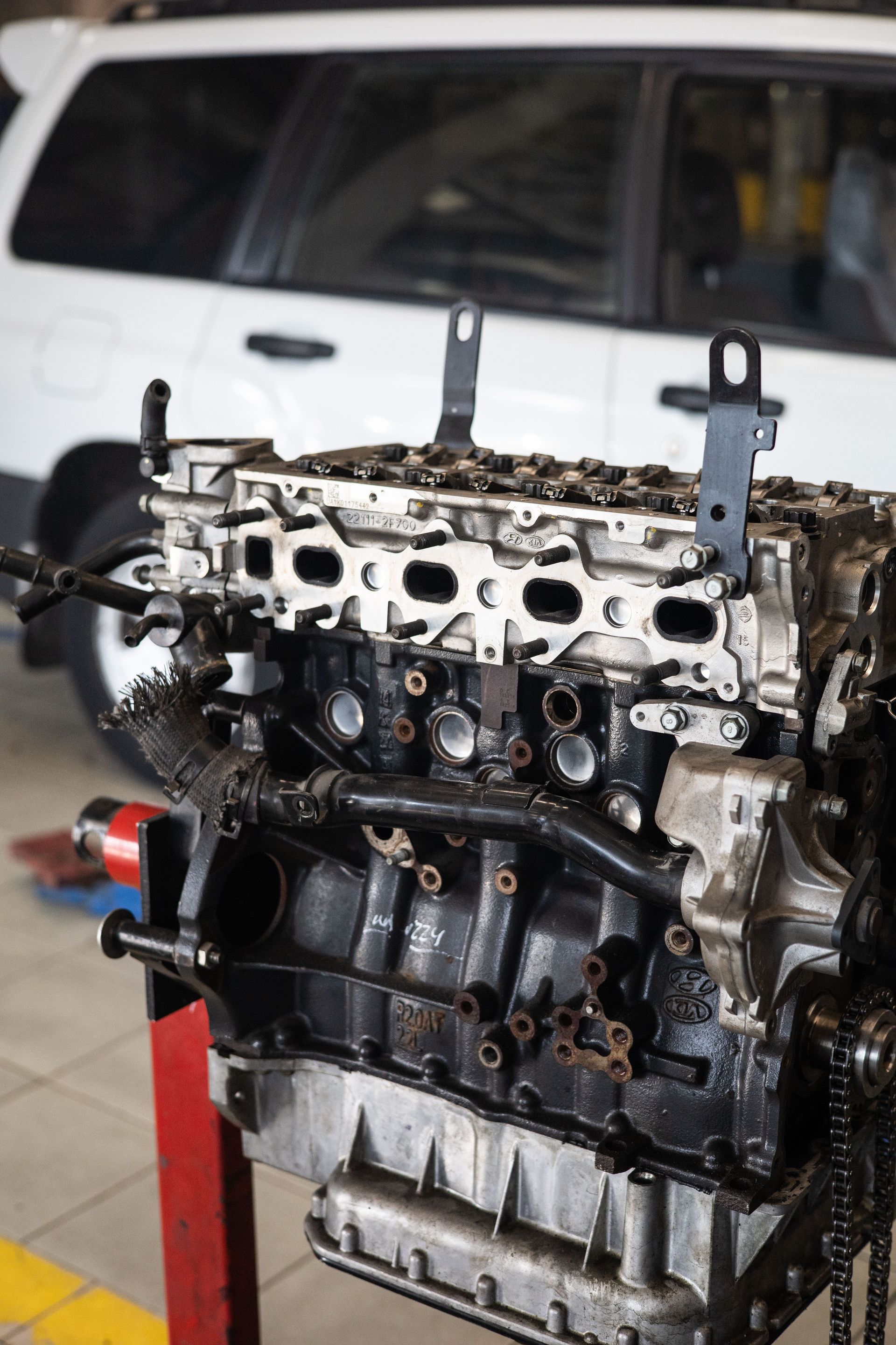 Exposed car engine block mounted on a stand, with a white vehicle in the background.