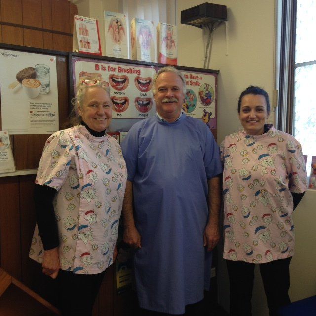 Three restaurant staff members in patterned shirts stand beside a wall of dessert posters.
