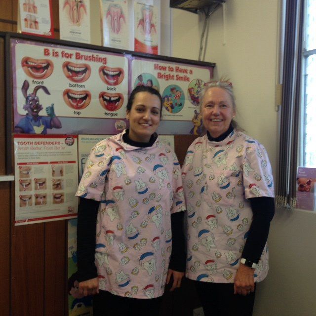 Two women in pink dental bibs stand by a brushing-and-flossing poster.
