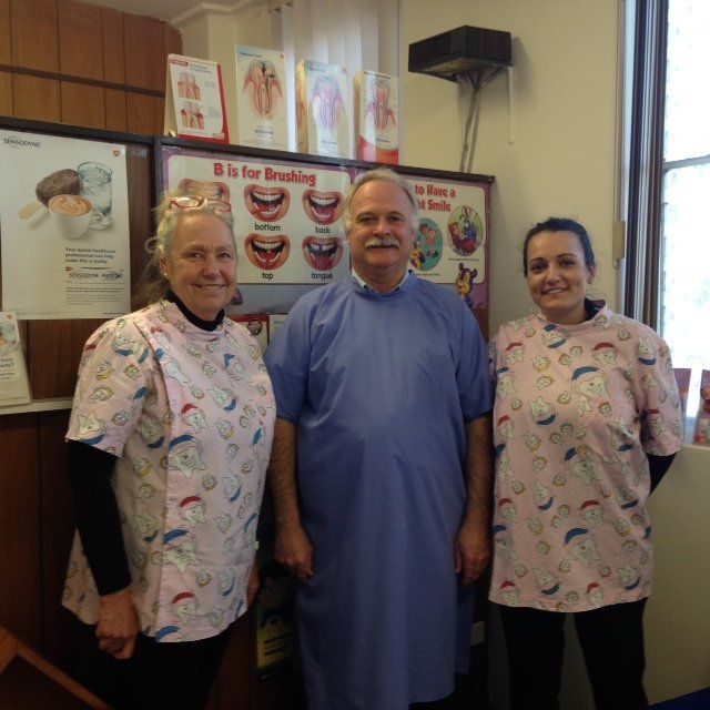 Three people in pastel smocks posing in a casual indoor setting, smiling beside a wall of colorful posters.