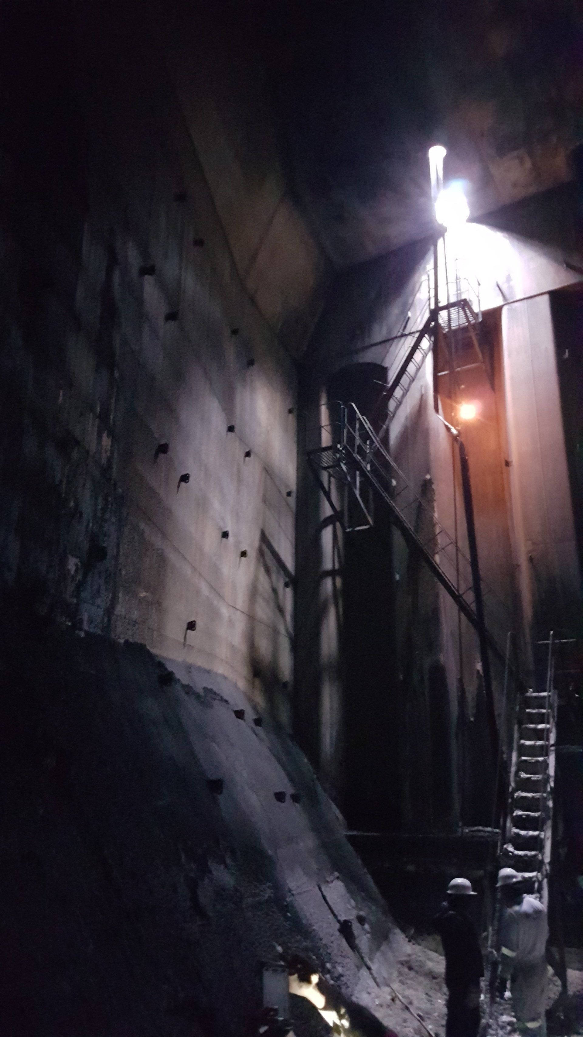 Donkere industriële ruimte met zichtbare rotsen, bouten en een ladder. Heldere plafondlampen verlichten het tafereel.