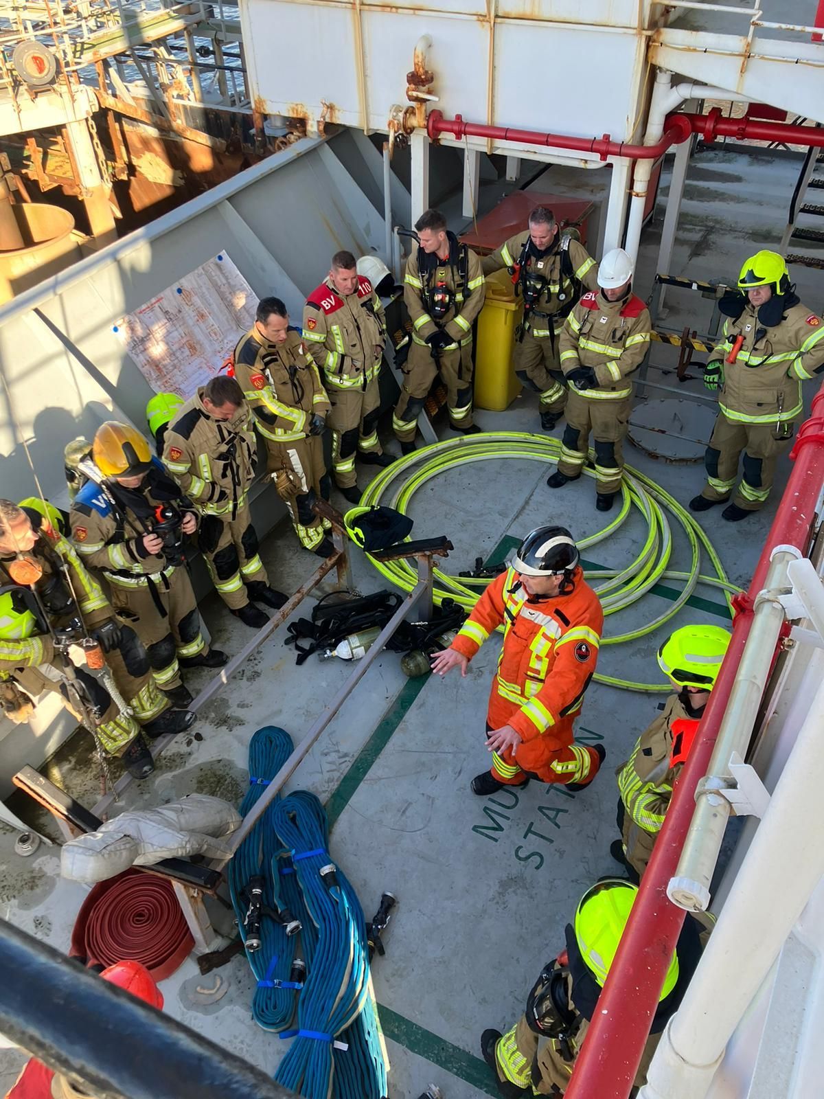 Brandweerlieden in uniform op een dek luisteren naar een instructeur. Gele slangen.