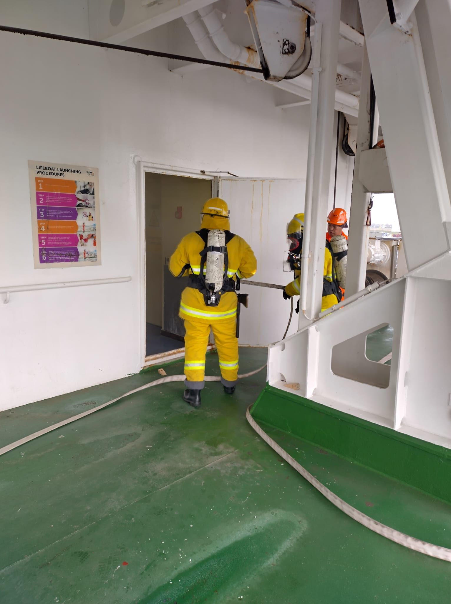 Brandweerlieden in gele pakken en met ademhalingsmaskers gaan een deuropening van een schip binnen.