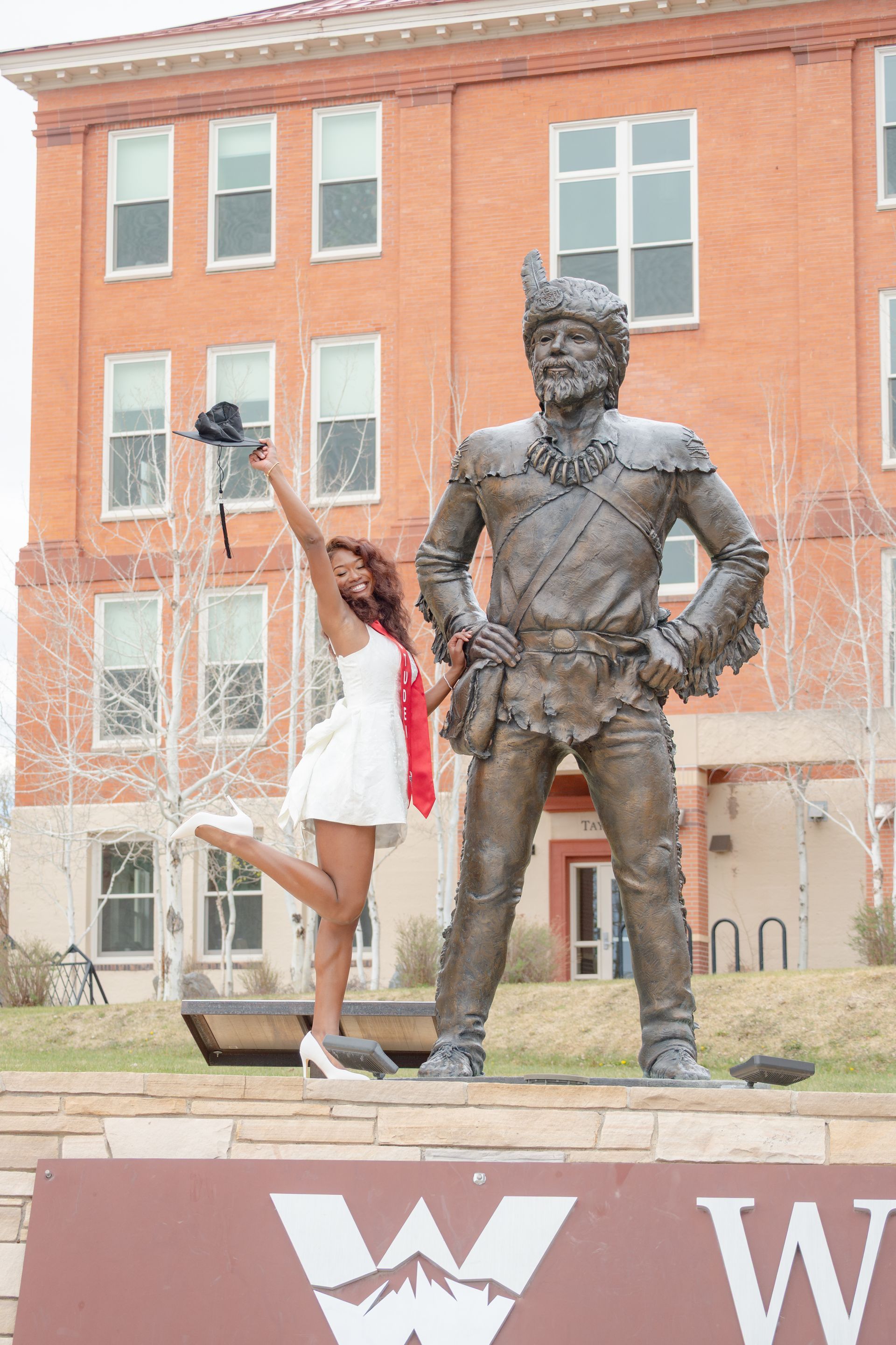 Person posing beside a bronze statue in front of a brick building, holding up a phone on a selfie stick.