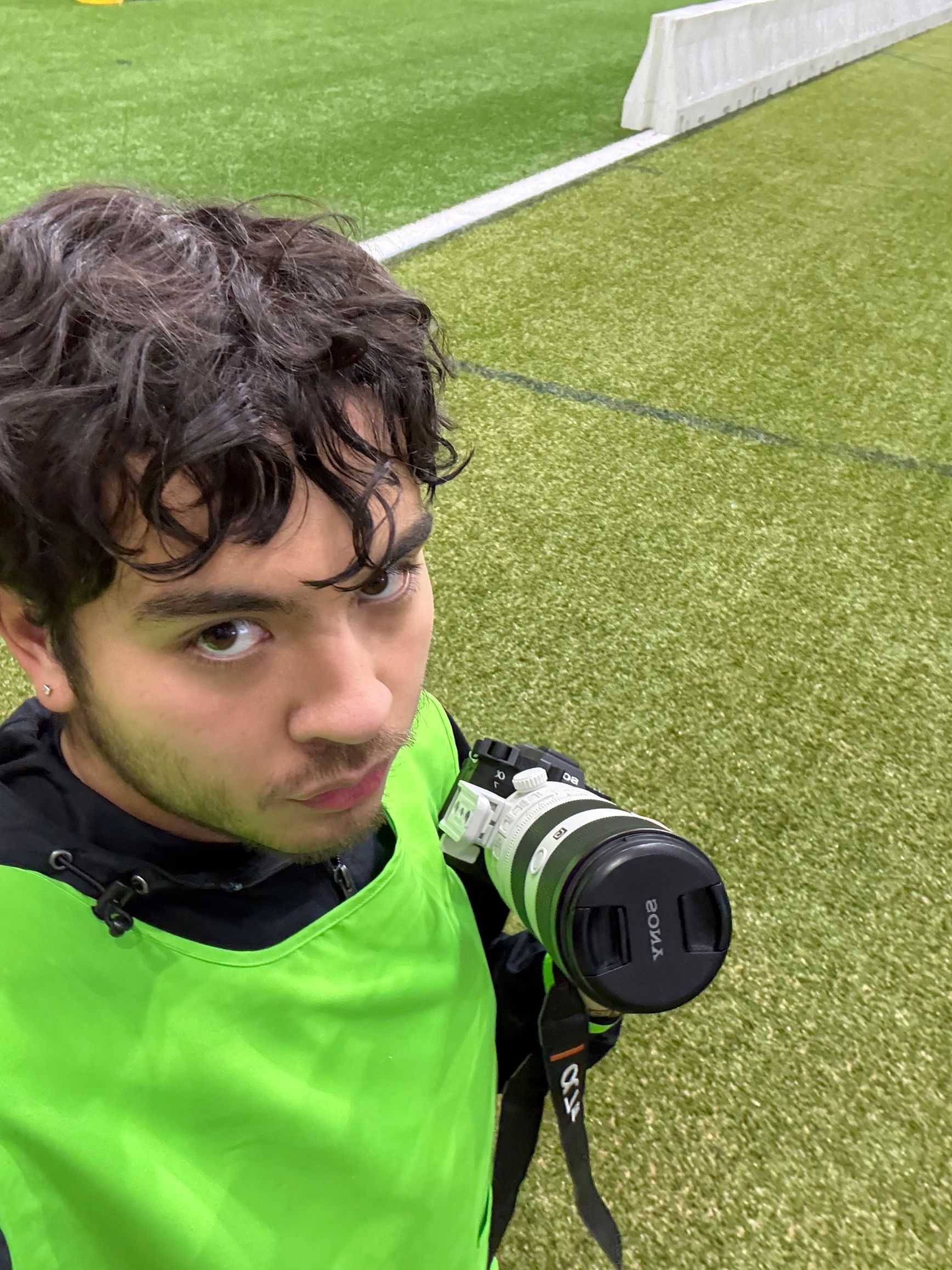 Kai Vong in a neon green vest holding a camera on a soccer field.