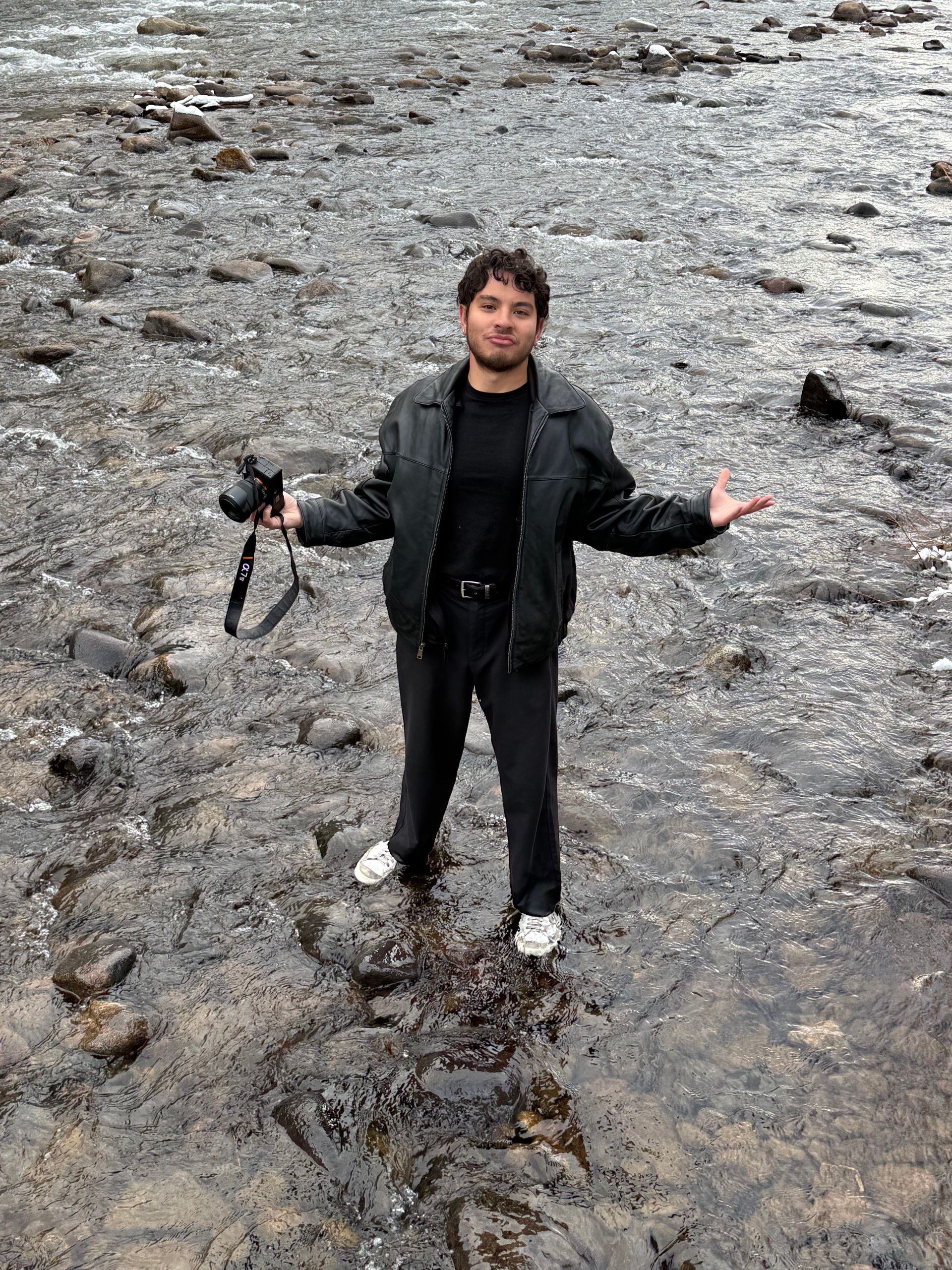 Kai Vong standing on a rocky shore, holding a camera and gesturing with open arms.