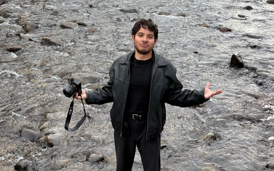 Kai Vong standing on a rocky shore, holding a camera and gesturing with open arms.
