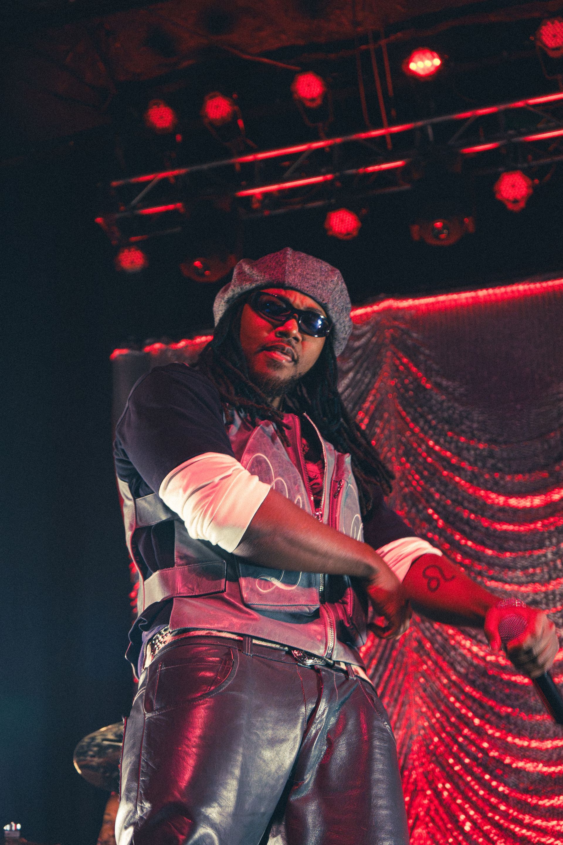 Leon Thomas in black leather outfit and hood on a red-lit stage, posing in front of draped curtains