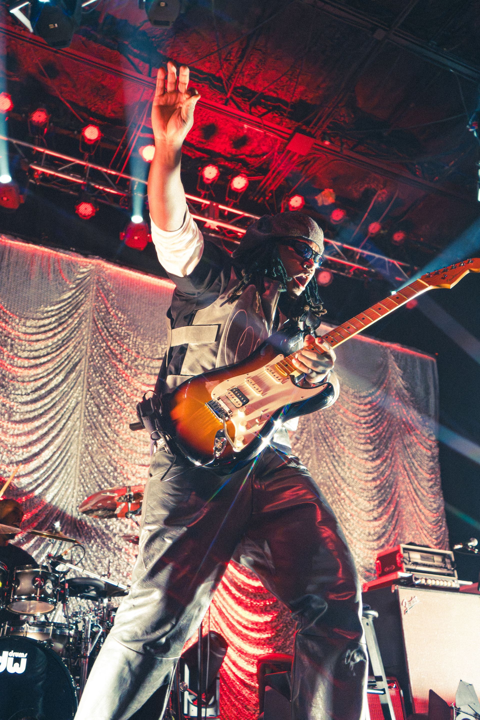 Leon Thomas on stage under red lights, raising one arm while playing an electric guitar