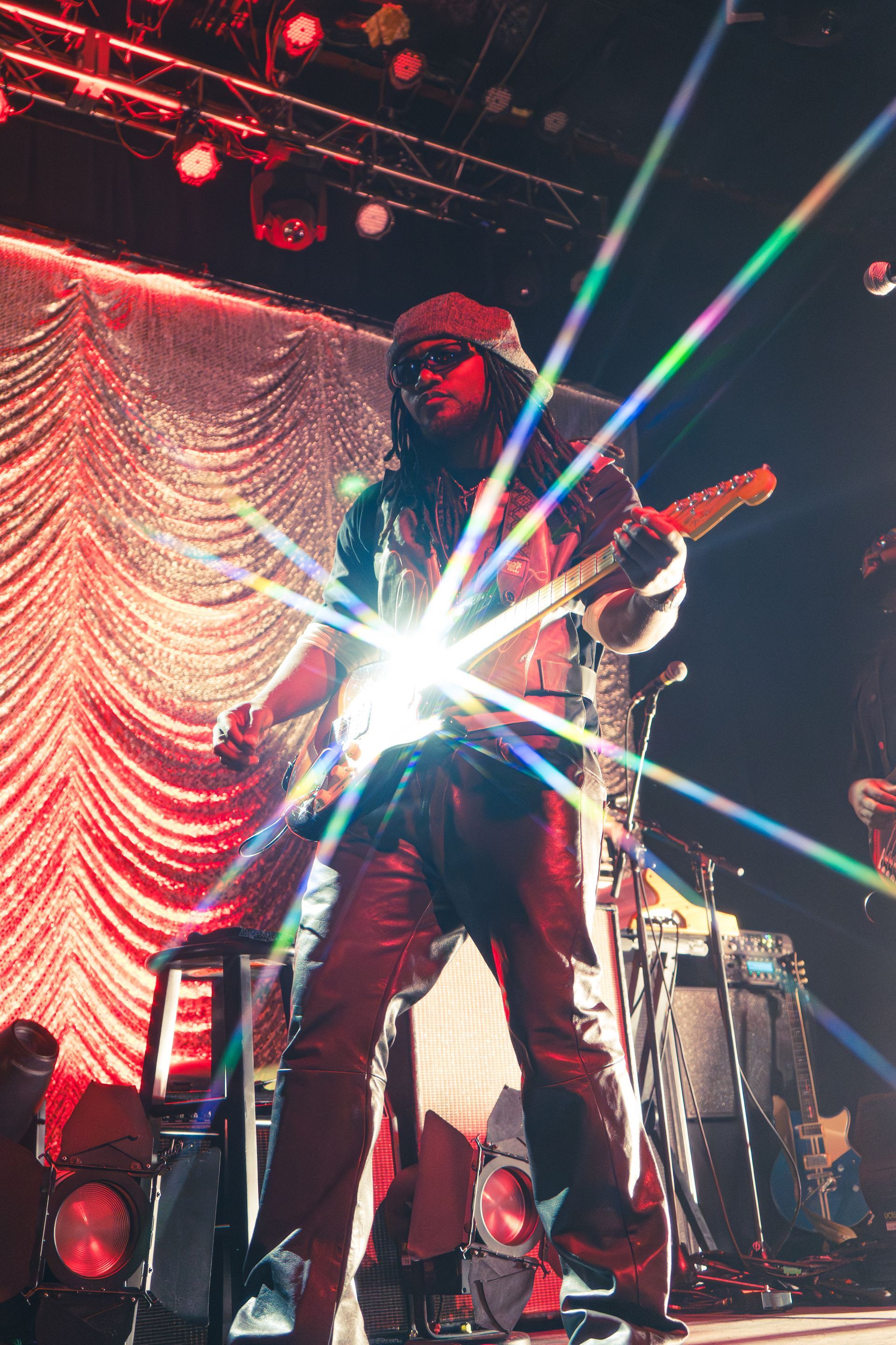Leon Thomas performing on stage under red lights, with bright starburst from the guitar and drum kit behind.