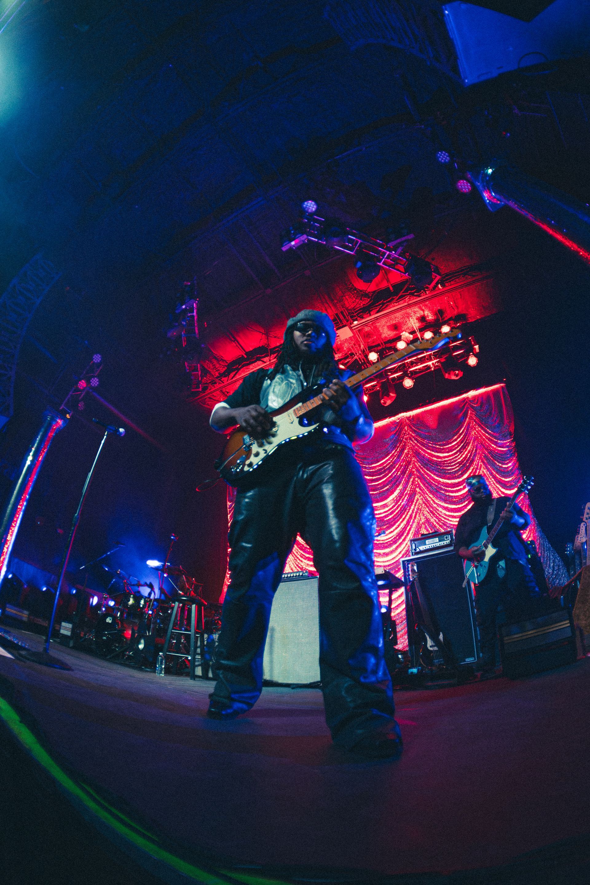 Leon Thomas playing guitar on a dim stage with red and blue lighting and a large backdrop