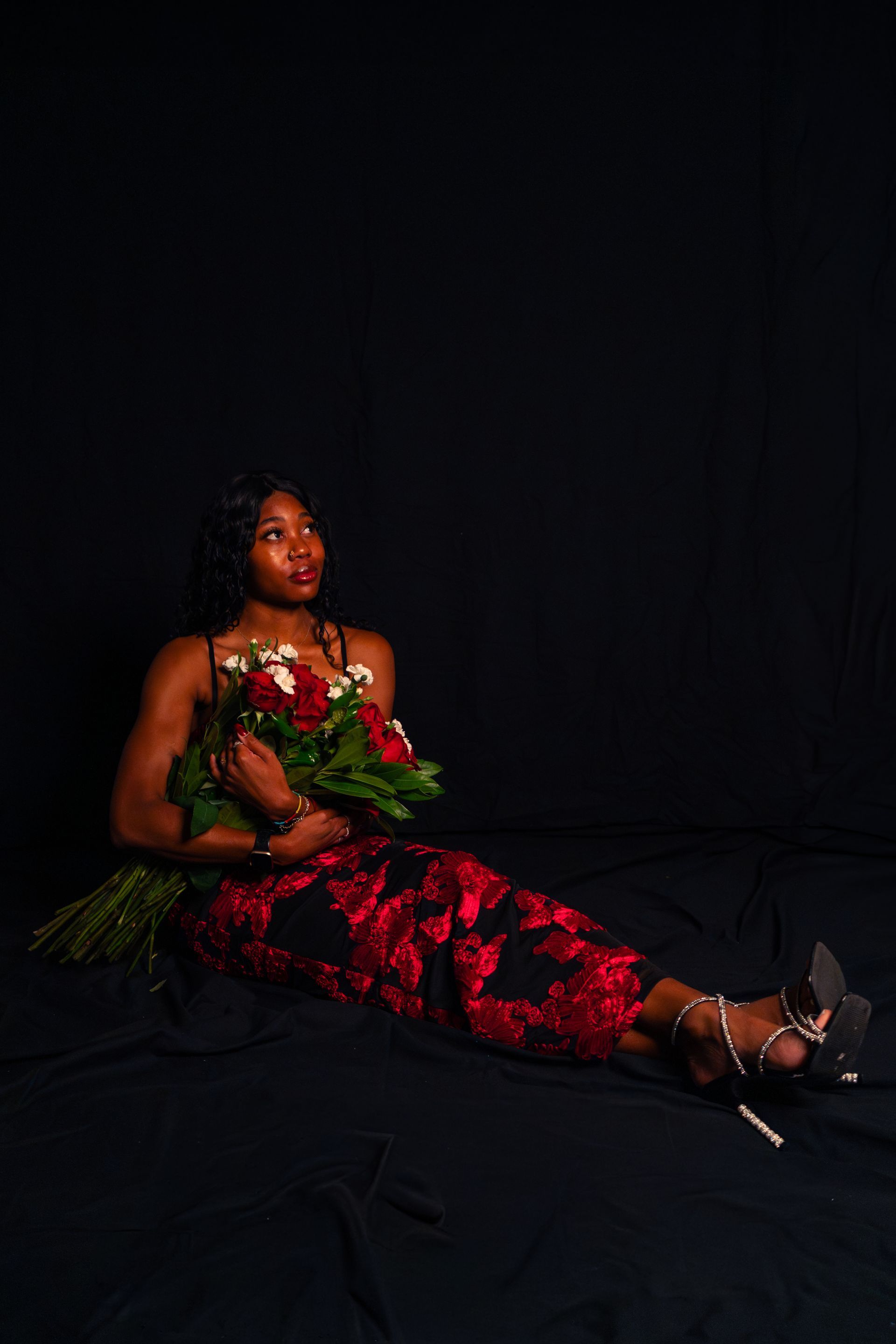 Woman in a red floral dress seated on a dark stage, holding flowers and looking upward.
