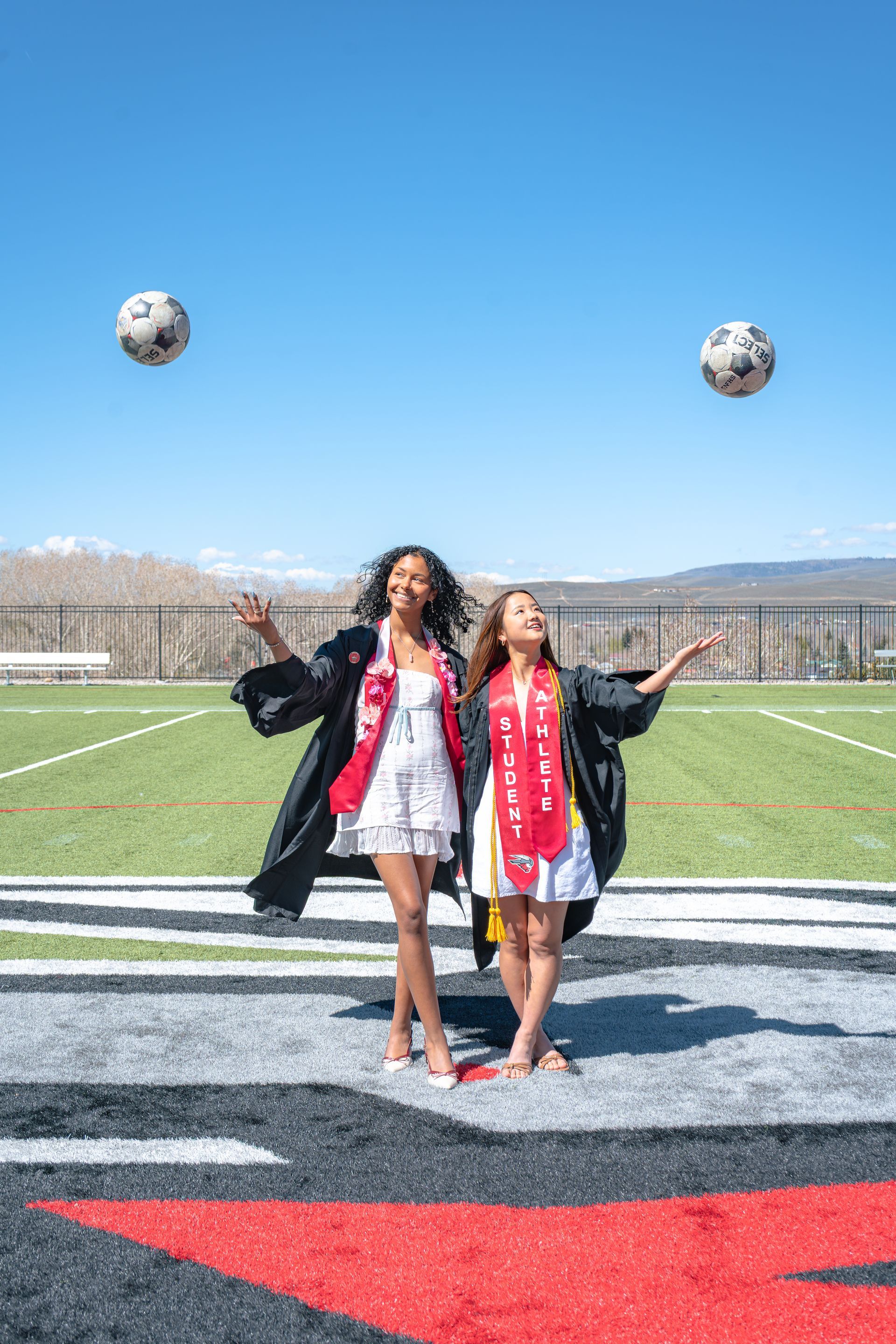 Two graduates in caps and gowns pose on a football field with soccer balls in the air.