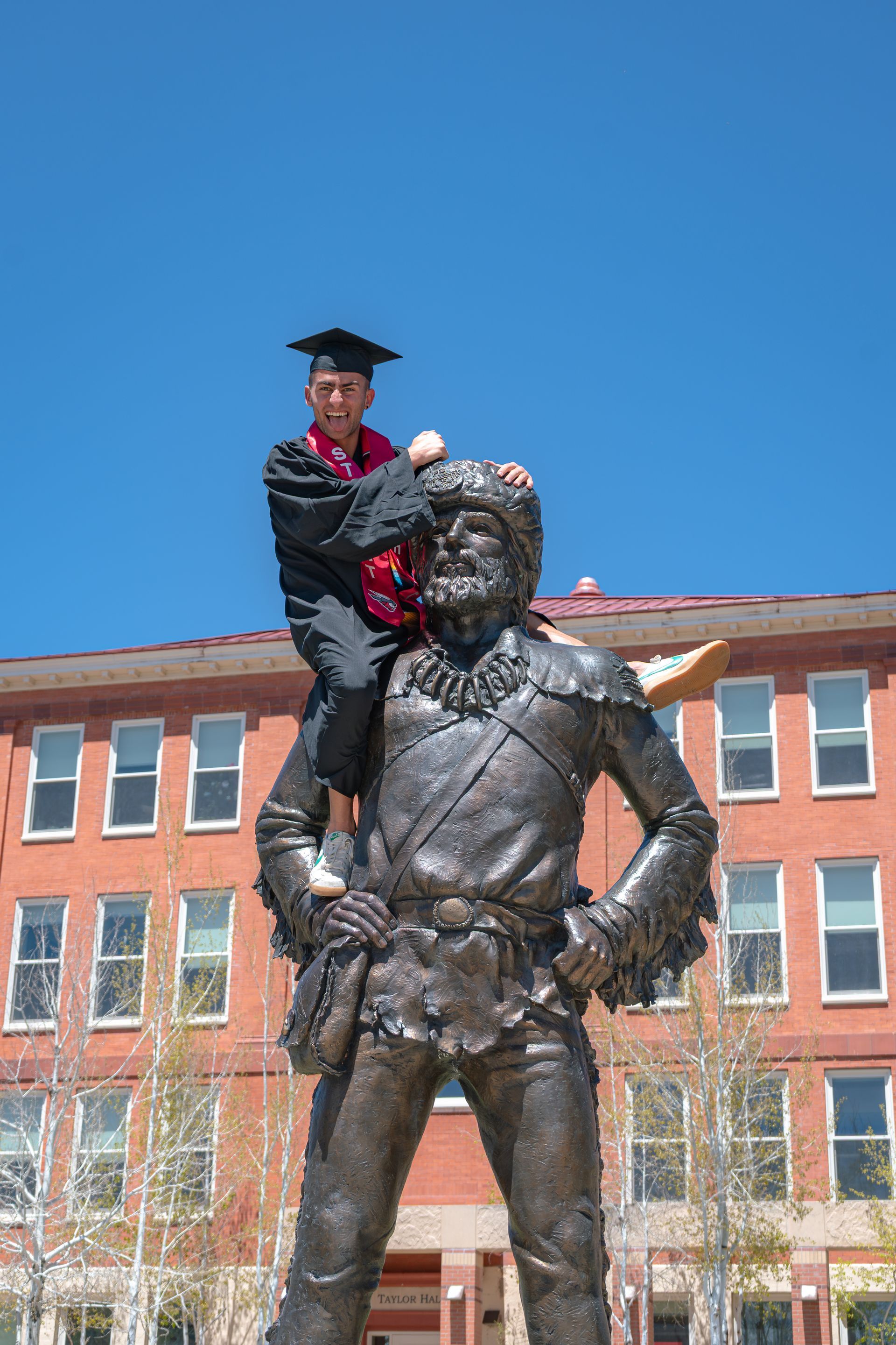 Person in red hat perched on a bronze statue in front of a brick building under a blue sky