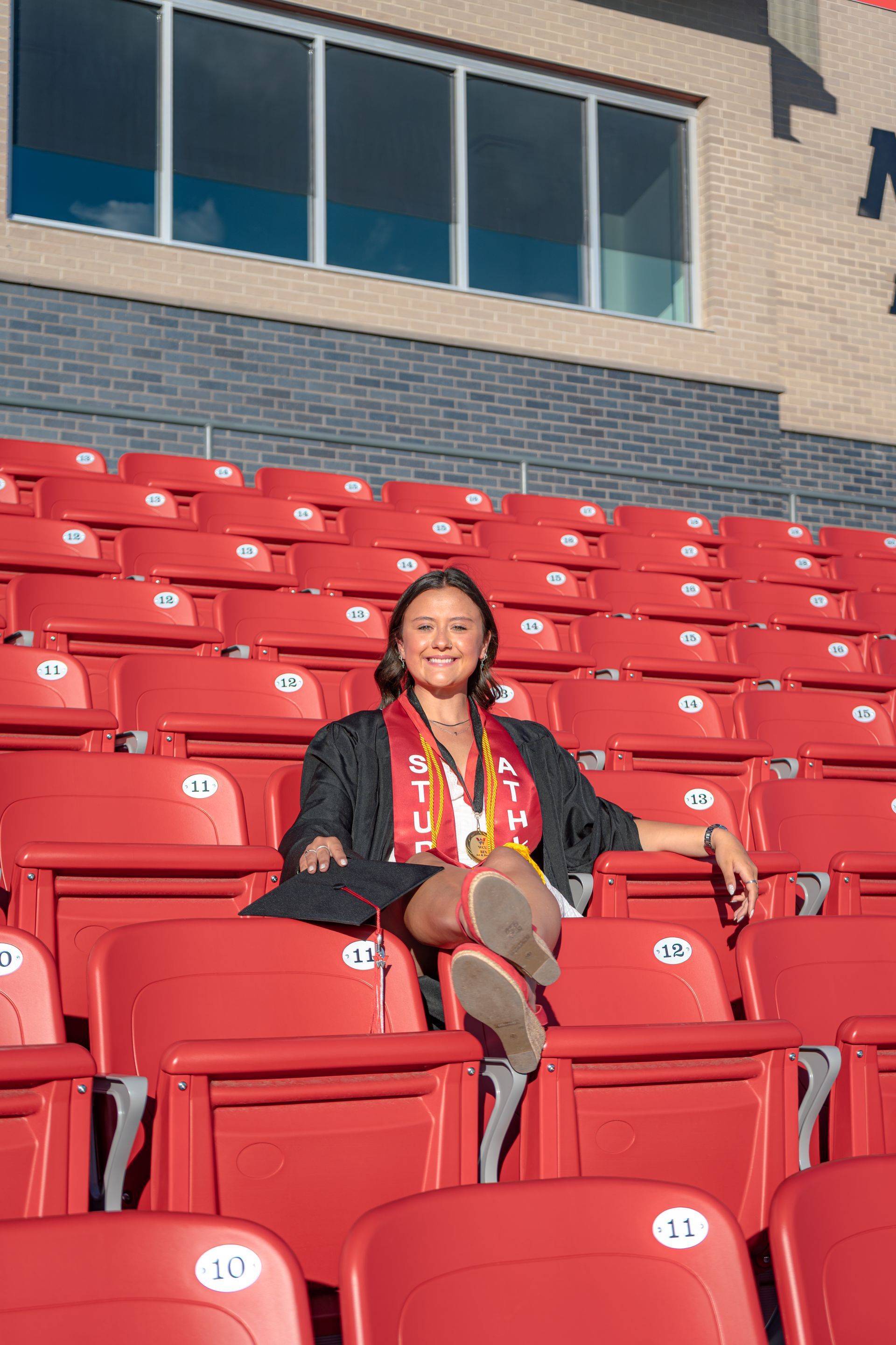 Person seated among red stadium seats, wearing a black jacket and colorful scarf, in front of a building.