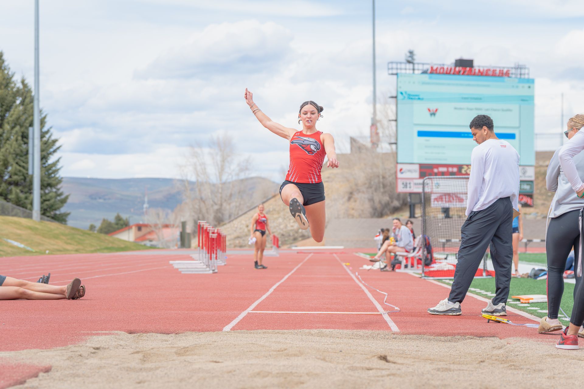 Runner mid-jump over a track sand pit during a long jump event, with spectators nearby.