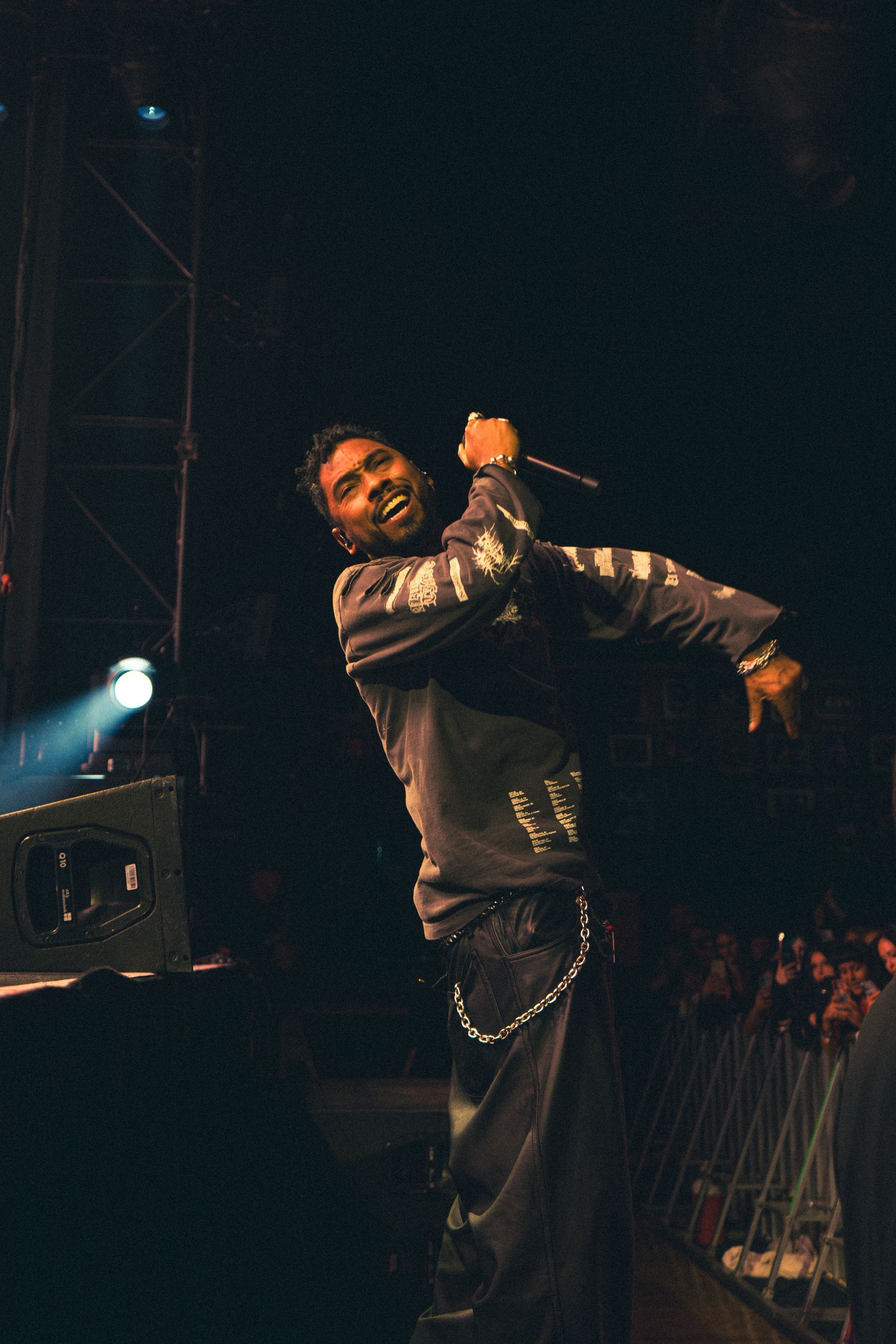 Miguel in a black jacket performing on a dark stage with a microphone, arm raised.