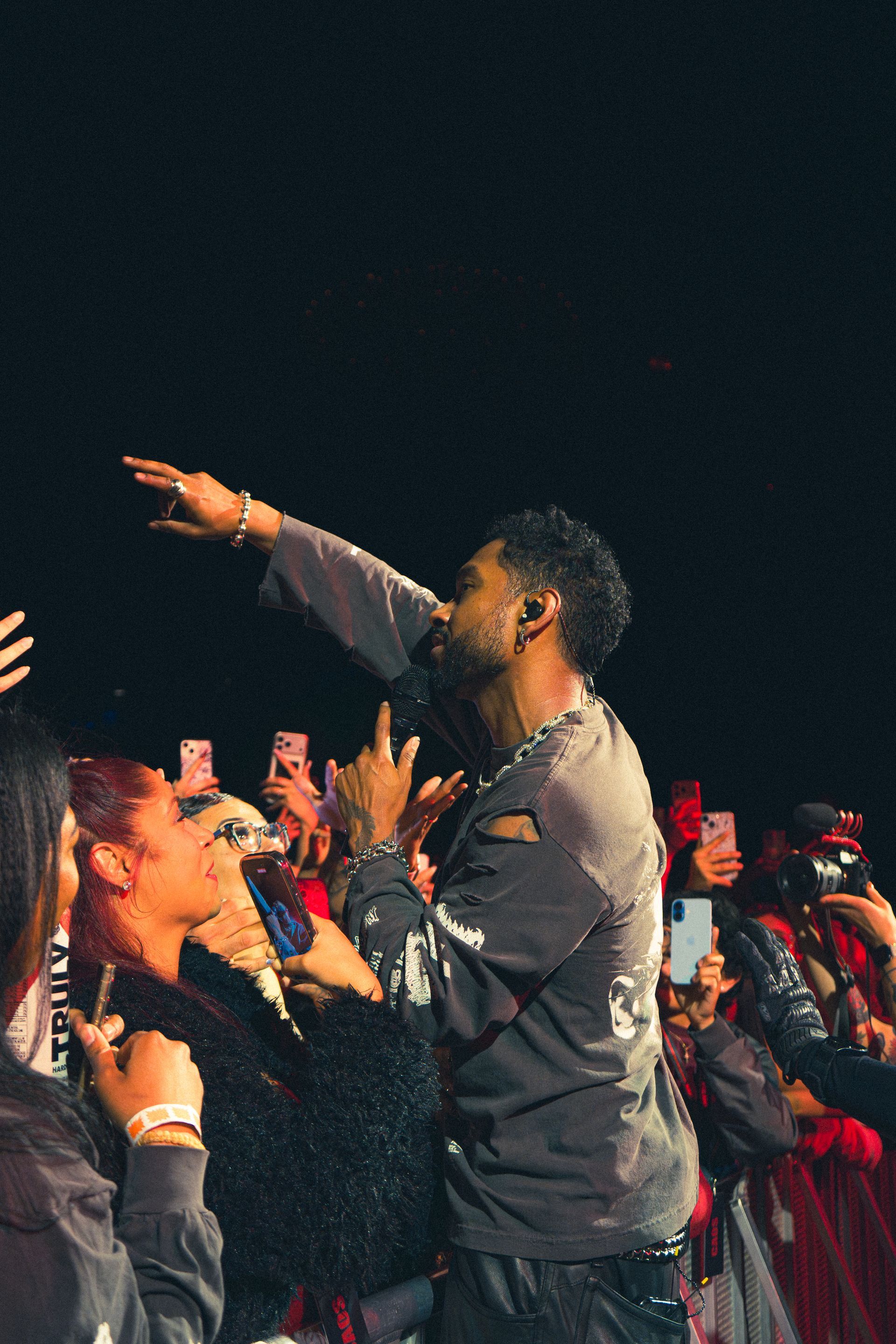 Miguel reaching toward cheering crowd at a concert, with raised hands and stage lighting.