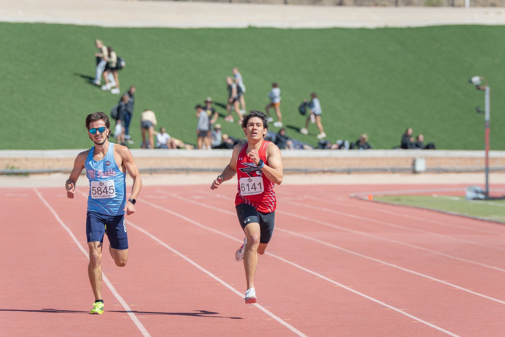 Two runners sprint on a track, with spectators and a grassy field in the background.