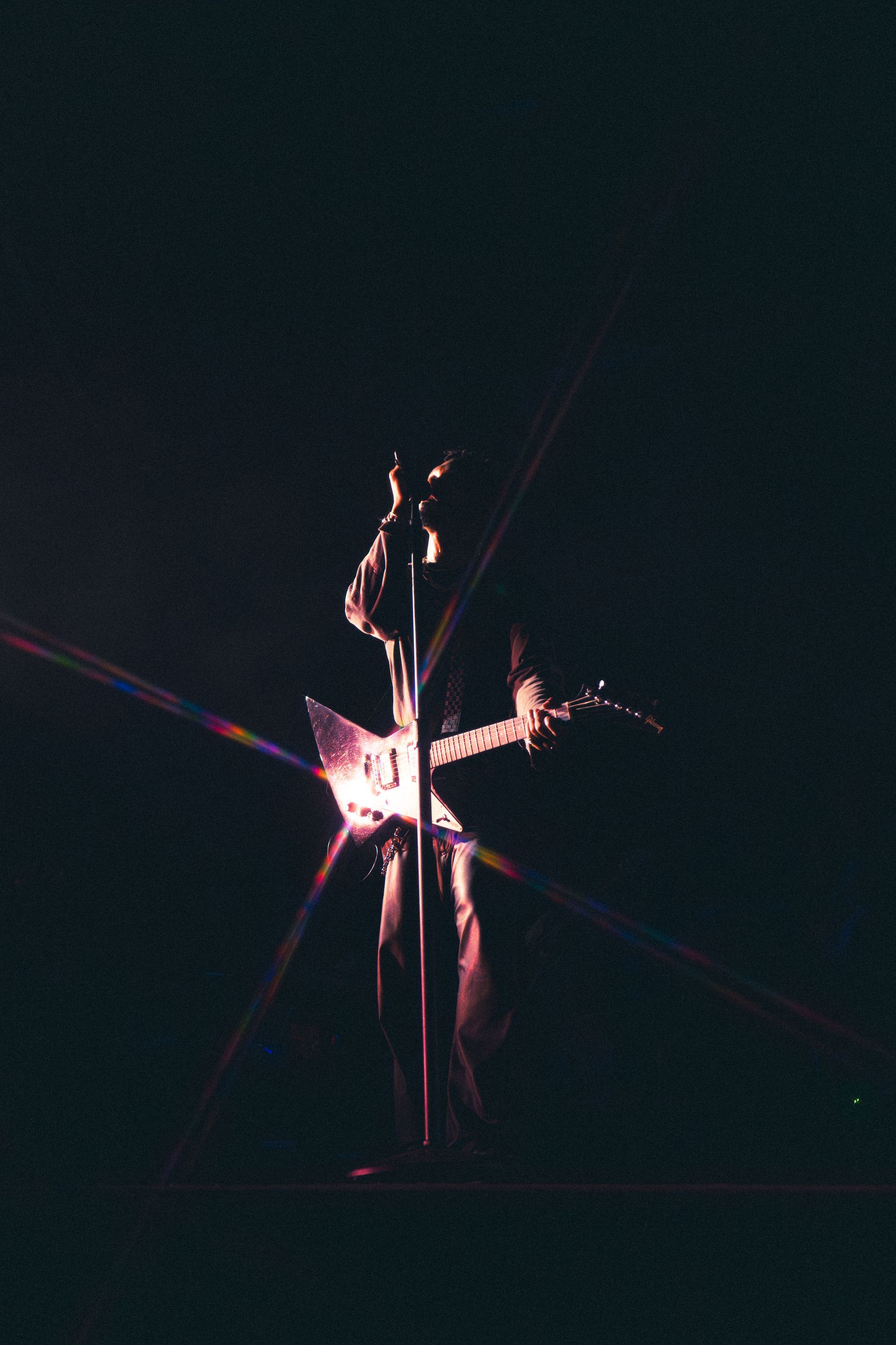 Dim stage silhouette of Miguel holding a guitar, lit by pink and white spotlights.