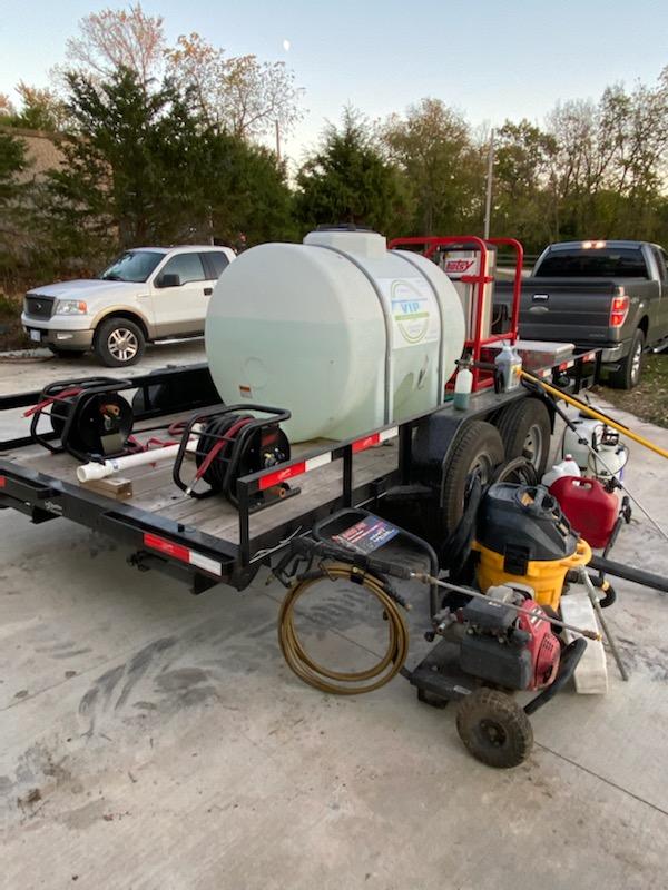 Trailer with water tank and equipment, parked on a concrete surface, with a white truck on the left and a black truck on the right.