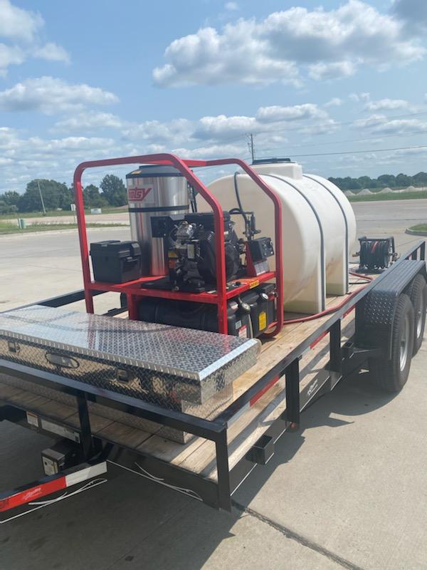 Pressure washing equipment on a trailer; red cage, white tank, silver toolbox, blue sky.
