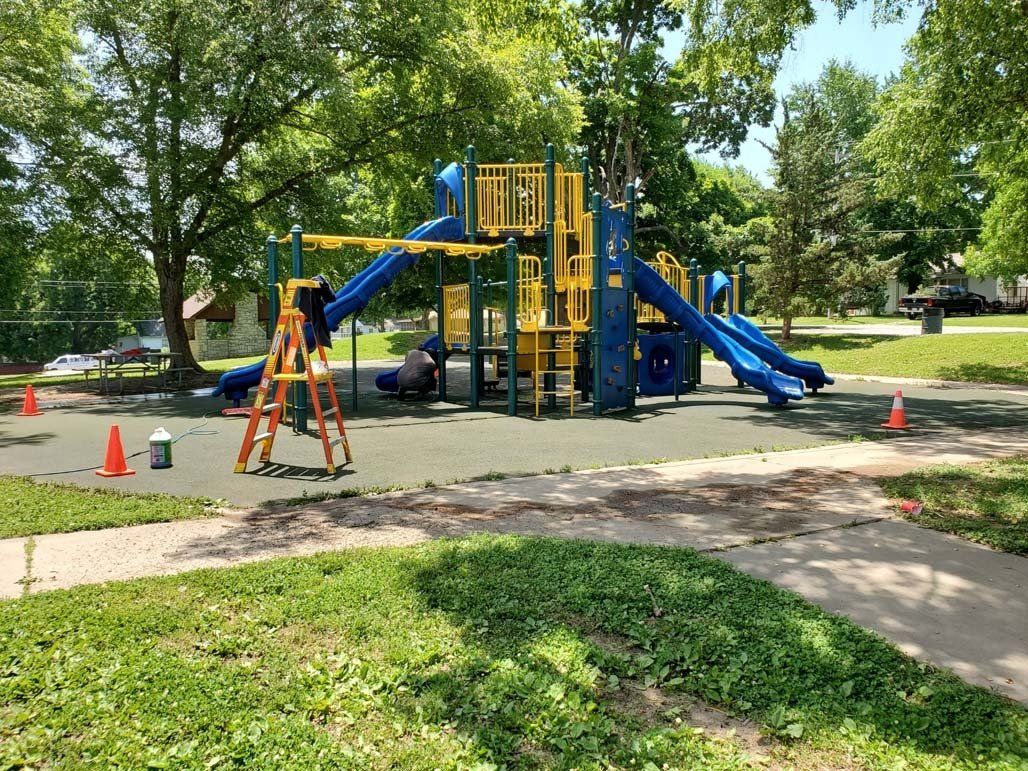 Playground with blue and yellow equipment, orange cones, and a ladder; workers are present.