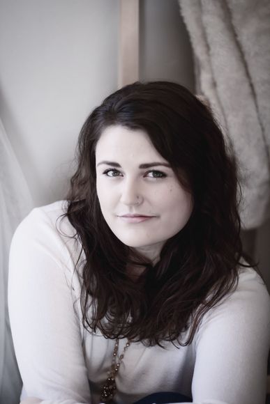 A woman with long brown hair is sitting in front of a window and looking at the camera.