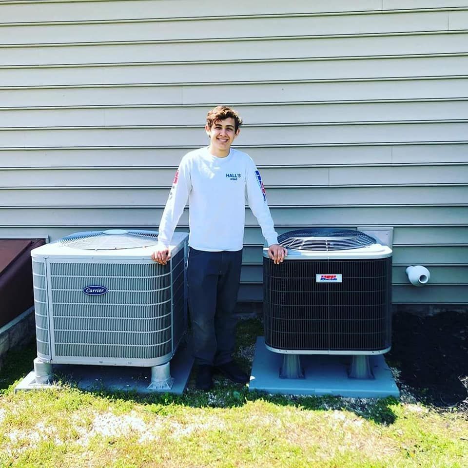 Young person stands between two air conditioning units. Outdoors, gray siding, and grass.