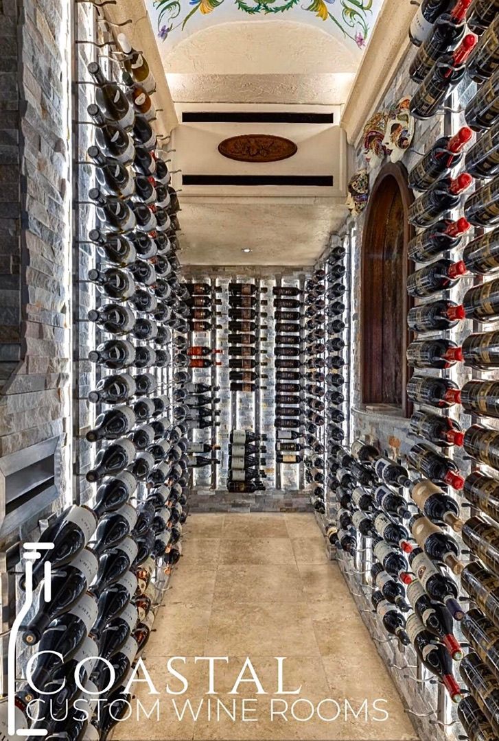 A narrow wine cellar with floor-to-ceiling metal racks holding numerous wine bottles, featuring a stone floor and archway.