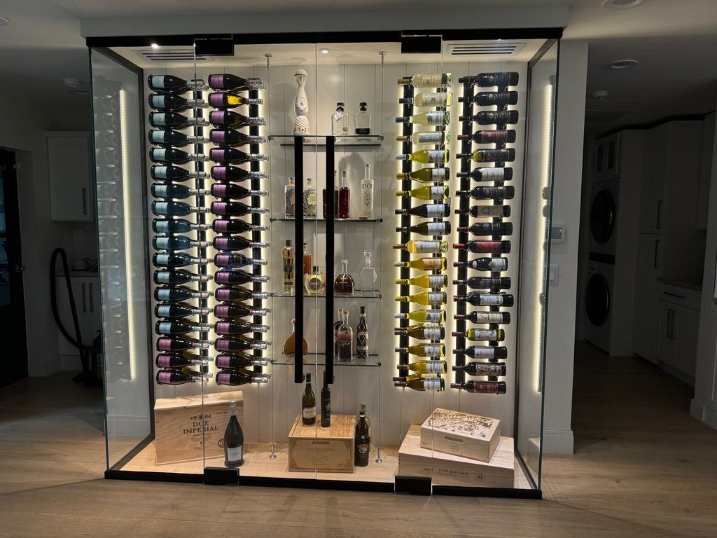 A lit, modern glass wine display featuring two vertical bottle racks on the sides and center shelves for spirits.