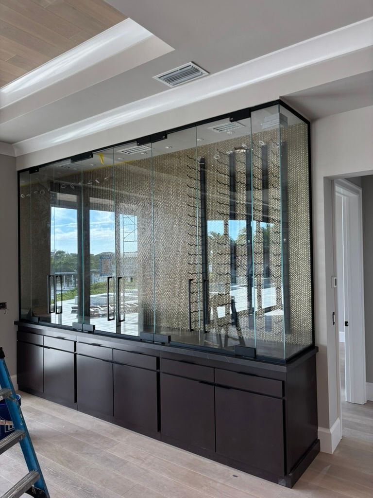 A custom glass wine cabinet with metal racks sits atop a dark wood base in a modern, light-filled room.