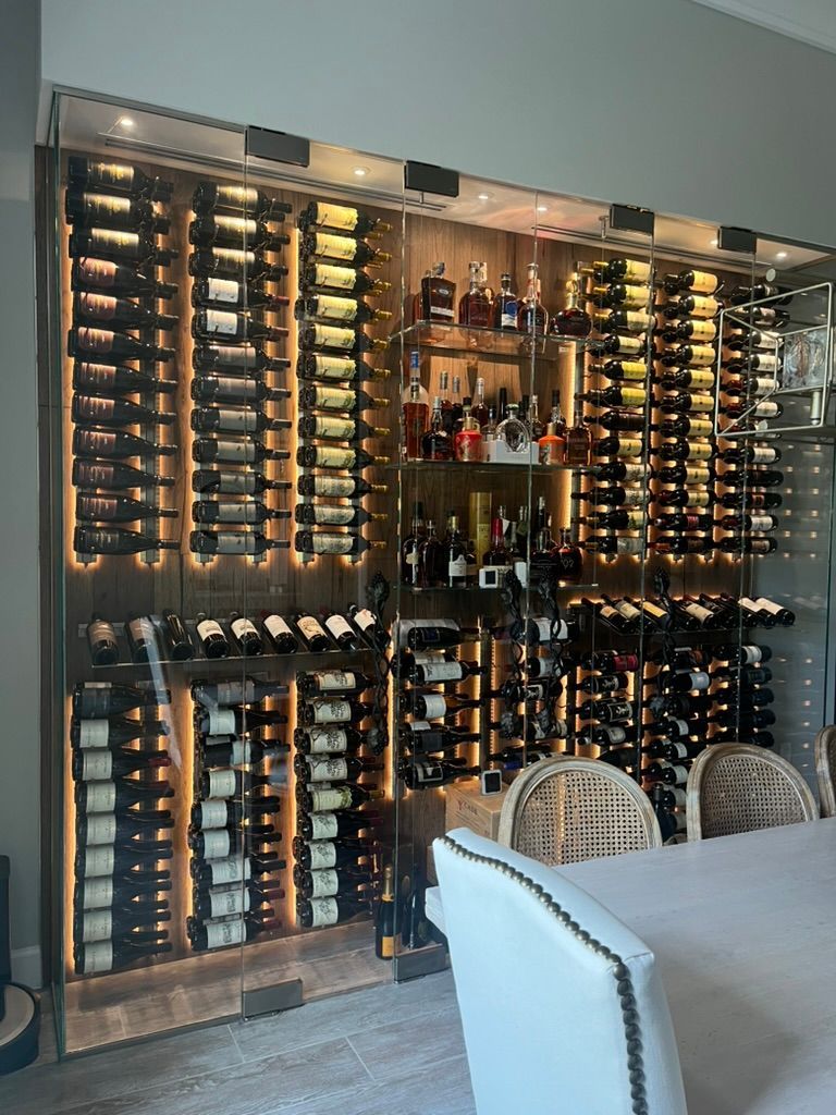 A large, floor-to-ceiling illuminated wine rack filled with many bottles, positioned behind a dining table and chairs.