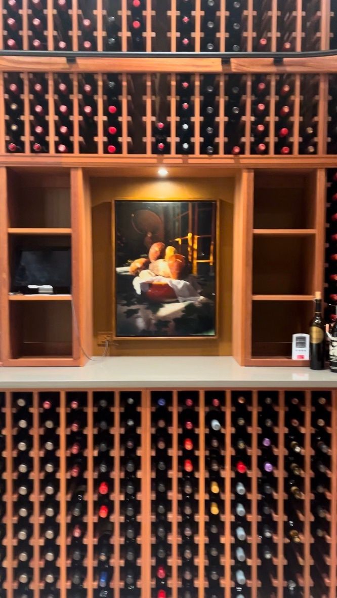 A wooden wine rack wall featuring a central alcove with a framed painting, illuminated by a small overhead light.