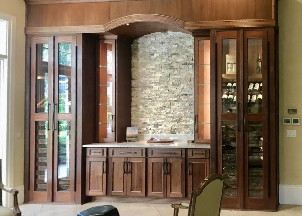 A built-in dark wood wine bar with glass-front cabinets flanking a central stone wall, arched top, and white countertop.