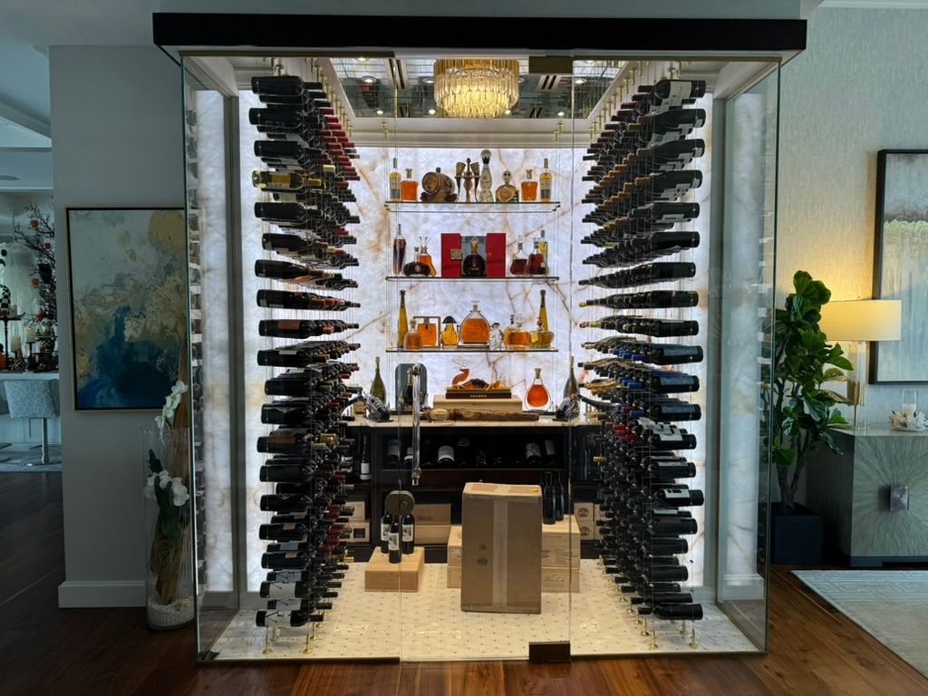 A glass-enclosed wine cellar with wall-mounted racks, illuminated marble backlighting, and shelves of liquor bottles.