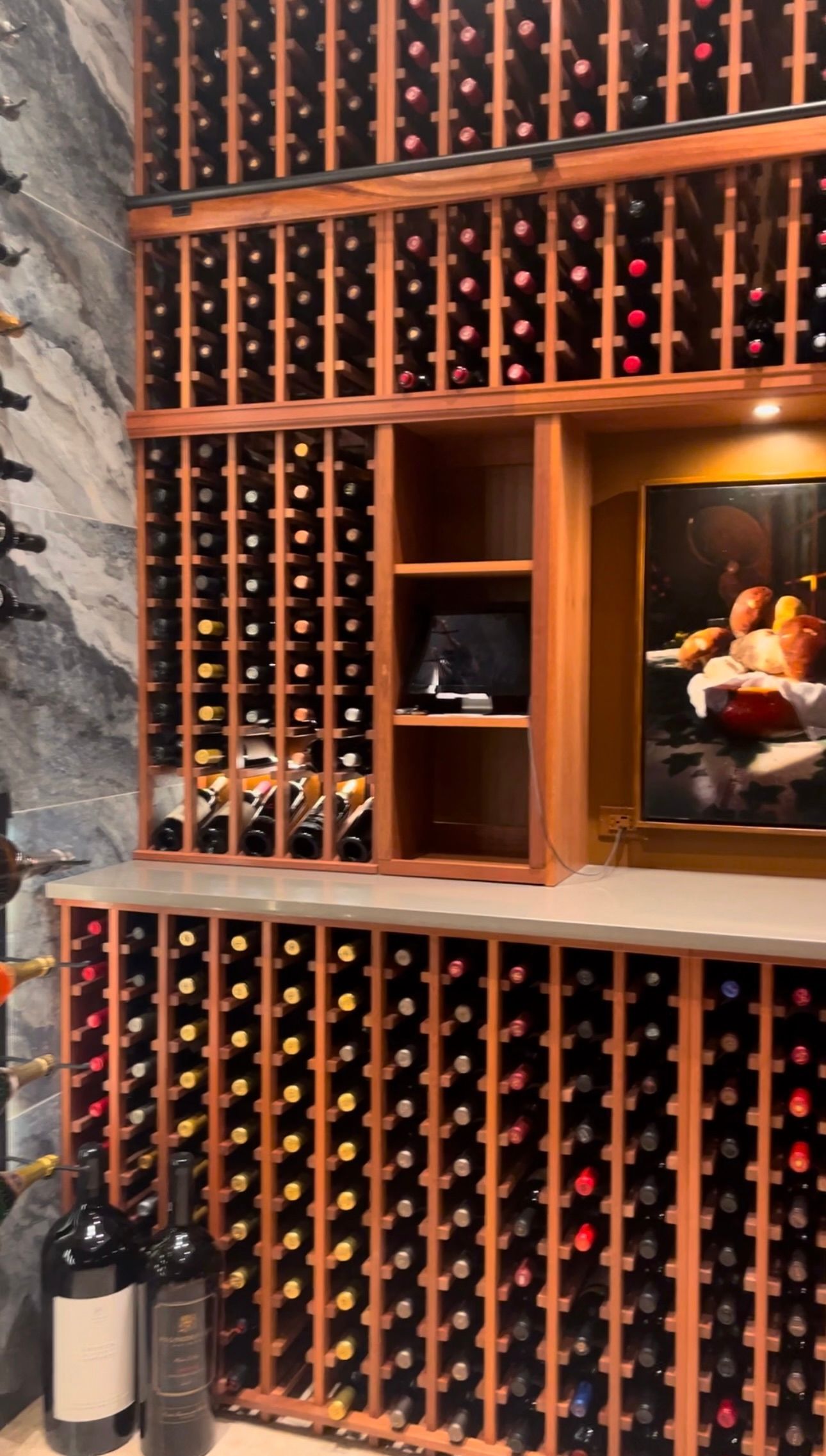 A wooden wine rack filled with bottles, featuring a central display shelf and a framed still-life painting.